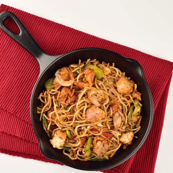 Chicken and shrimp noodles with vegetables in a black skillet on a red placemat.