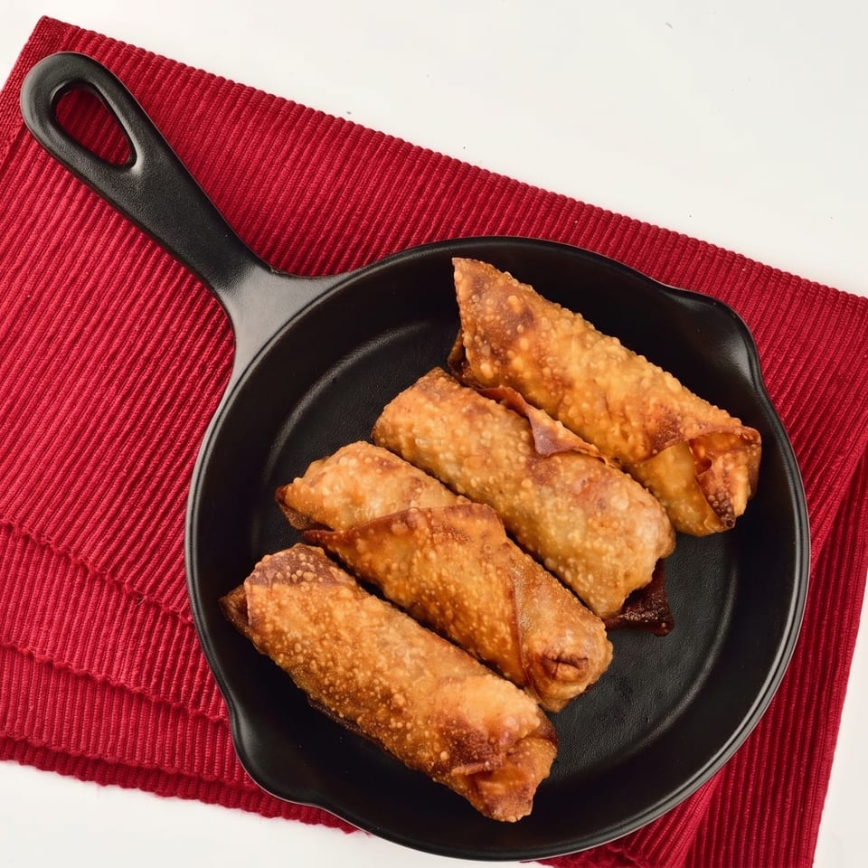 Four crispy, golden-brown egg rolls arranged in a black skillet on a red placemat.