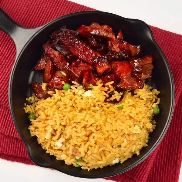 A black skillet holds glistening red char siu pork and golden fried rice with peas.