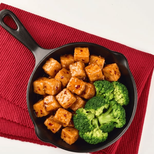 Sweet and sour tofu with broccoli florets in a black skillet on a red placemat.